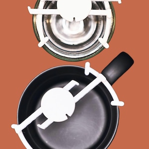 May include: Overhead view of two cooking vessels with white trivets. The top vessel is a clear glass jar, and the bottom is a black pan with a handle. Both have white trivets with a circular centre and four arms.