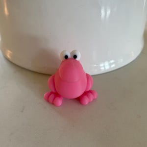 May include: A small, pink frog figurine with large, white-and-black eyes. The frog has four webbed feet and a smiling expression. The figurine is sitting on a light-colored surface, with a white object in the background.
