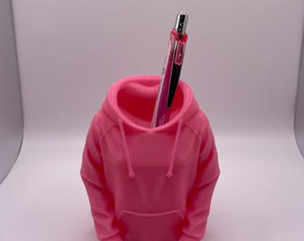 May include: A vibrant pink pen holder shaped like a hoodie. The pen holder has a hood, a front pocket, and drawstrings. A pen with a black and pink barrel is inside the pen holder. The pen holder is made of a smooth material.