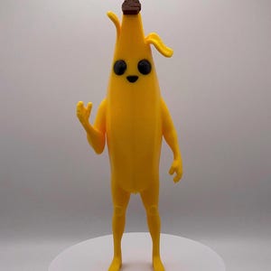 May include: A yellow banana-shaped figurine with black eyes and a smiling mouth. The figurine has arms and legs, and a brown top. It is standing on a white surface.
