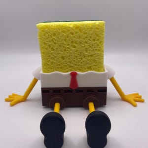 May include: A novelty sponge holder shaped like a cartoon character. The holder has a yellow sponge head, white shirt, brown pants, yellow arms and legs, a red tie, and black shoes. The arms and legs are outstretched.