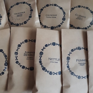 May include: A set of ten brown paper bags with black and white labels. Each bag has a different herb name and Latin name printed on it. The herbs include Calendula, Coltsfoot, Cloves, Juniper Berries, Nettle, and Fennel.