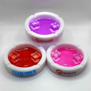 May include: Three white ceramic ashtrays with different colored liquids and ice cubes inside. The ashtrays have the words "WOCKHARDT", "Hi-Tech", and "QUAGEN" printed on the bottom.