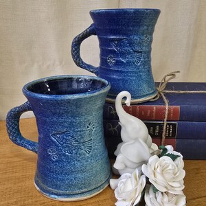 May include: Two blue ceramic mugs with textured handles and fish designs. A white elephant figurine and a bouquet of white roses sit near the mugs. The mugs are on a wooden surface with a stack of blue books in the background.