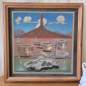 May include: Framed sand art depicting a desert landscape with a mountain, blue sky, and clouds. The artwork features several pottery vessels and a cow skull in the foreground. The frame is made of light brown wood.
