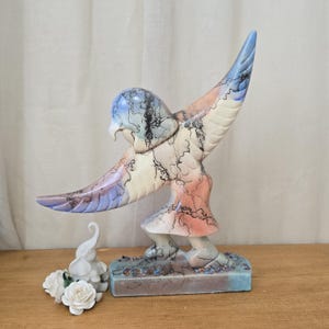 May include: A ceramic sculpture of a bird with outstretched wings, featuring a color palette of blue, pink, and cream. The bird is perched on a rectangular base. A small white elephant figurine and white roses are in the foreground.