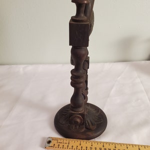 Op de afbeelding: Een donkerbruine, houten decoratieve standaard met een ronde basis en sierlijke gravures. De standaard heeft een centrale kolom met decoratieve elementen en een vierkante bovenkant. Een liniaal staat onderaan.