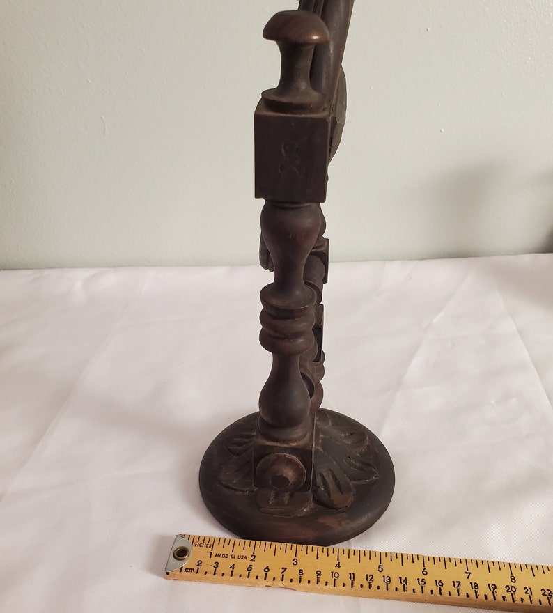Op de afbeelding: Een donkerbruine, houten decoratieve standaard met een ronde basis en sierlijke gravures. De standaard heeft een centrale kolom met decoratieve elementen en een vierkante bovenkant. Een liniaal staat onderaan.