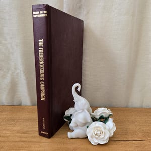 May include: A hardcover book titled "The Fredericksburg Campaign" with gold lettering, next to a white ceramic elephant figurine and white roses. The book has a dark burgundy cover.