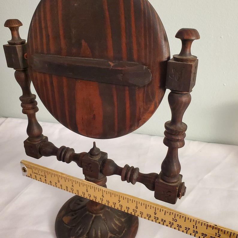 Op de afbeelding: Een vintage houten toilettafelspiegel met een ronde, donker gebeitste lijst en sierlijke details. De spiegel wordt ondersteund door een decoratieve voet en twee staanders. De lijst van de spiegel heeft een diameter van ongeveer 25 cm.