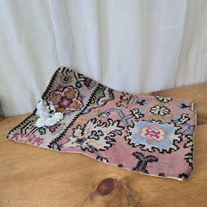May include: A rectangular, pink and patterned textile with a floral design, possibly a rug or decorative piece. White ceramic flowers and a swan figurine are placed on the textile. The textile is on a wooden surface.
