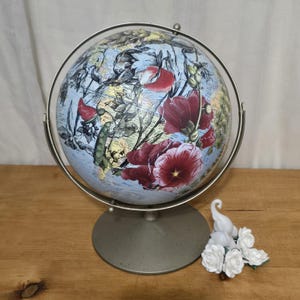 May include: A decorative globe with a floral design featuring red and pink flowers on a light blue background. The globe is mounted on a silver-colored metal stand. A small white elephant figurine and white roses are next to the globe.