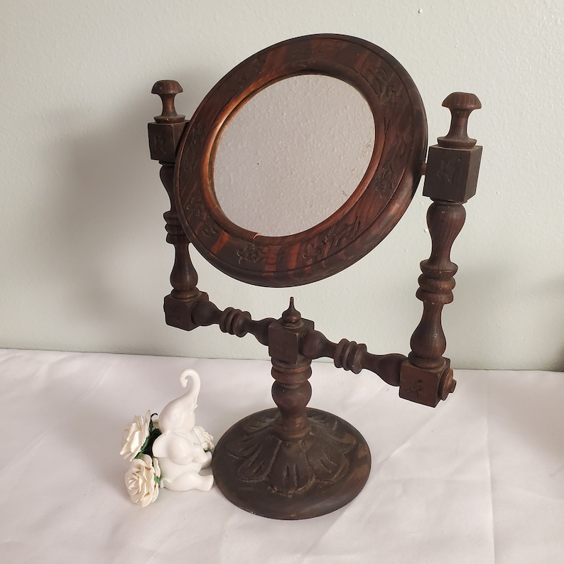 Op de afbeelding: Een vintage houten toilettafelspiegel met een ovale lijst en bloemsnijwerk. De spiegel wordt ondersteund door een gedetailleerde houten standaard. Het donkerbruine hout heeft een warme tint. Een klein wit olifantenbeeldje en een boeket witte bloemen staan op de voorgrond.