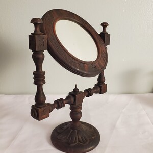 Op de afbeelding: Een antieke kaptafelspiegel van donkerbruin hout, met een ovale spiegel in een decoratief frame. De spiegel wordt ondersteund door een gedetailleerde houten standaard met een ronde basis en sierlijke gravures. De spiegel is iets gekanteld.