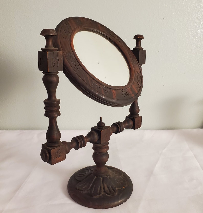 Op de afbeelding: Een antieke kaptafelspiegel van donkerbruin hout, met een ovale spiegel in een decoratief frame. De spiegel wordt ondersteund door een gedetailleerde houten standaard met een ronde basis en sierlijke gravures. De spiegel is iets gekanteld.