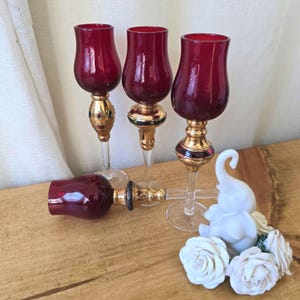 May include: Three red glass candle holders with gold accents and clear glass stems. The candle holders are arranged in a row on a wooden surface. A white ceramic elephant figurine is in the foreground.