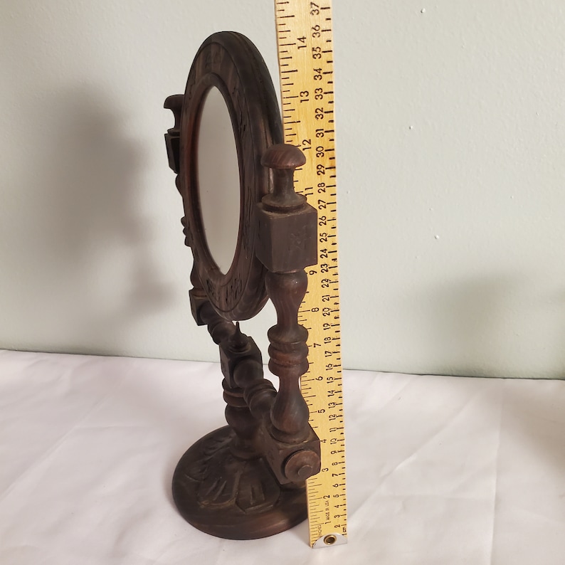 Op de afbeelding: Een donkerbruine make-upspiegel in antieke stijl. De ovale spiegel is omlijst door gesneden hout, ondersteund door een decoratieve standaard. De spiegel is ongeveer 30 cm hoog.