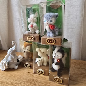 May include: Five small plush animal figurines displayed in clear plastic boxes with brown bases. The animals include a white rabbit, a gray cat, a fox, an elephant, and a skunk. Each box has the word "ARA" printed on the front.