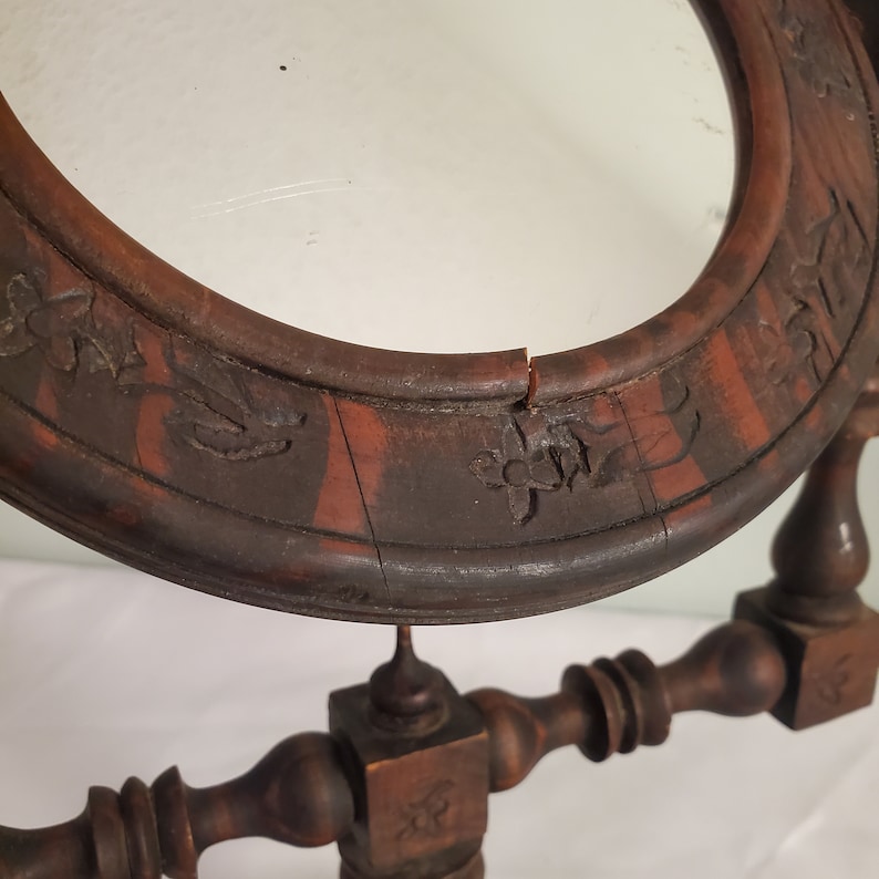 Op de afbeelding: Close-up van een vintage houten toilettafelspiegel. De ovale lijst is donkerbruin met gesneden bloemendetails en een scheur. De spiegel wordt ondersteund door een decoratieve houten standaard.