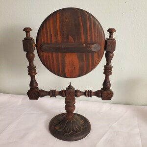 Op de afbeelding: Een vintage houten scheerspiegel. De ronde lijst is gemaakt van gebeitst hout, ondersteund door gedraaide houten palen en een decoratieve voet. Het hout heeft een roodbruine tint. De spiegel heeft een diameter van ongeveer 25 cm.