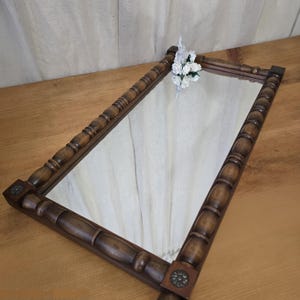 May include: A rectangular, wooden-framed mirror with a dark brown finish. The frame features a carved design with rounded and elongated elements. Small, decorative metal accents are at each corner. A small bouquet of white flowers rests on the mirror's surface.
