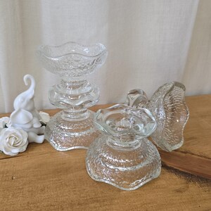 May include: Clear glass candle holders with textured surfaces and scalloped edges. The candle holders have a pedestal base and a bowl-shaped top. The image shows two complete candle holders and a third one that is partially visible.