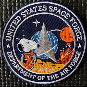 Może przedstawiać: Okrągła naszywka z białym napisem "UNITED STATES SPACE FORCE DEPARTMENT OF THE AIR FORCE" na granatowym tle. Naszywka zawiera Snoopy'ego w pomarańczowym skafandrze, srebrną gwiazdę, globus i pomarańczową planetę.