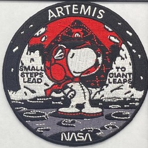 May include: Embroidered patch featuring Snoopy in a red and white spacesuit, with the word "ARTEMIS" at the top. The patch includes the NASA logo and the phrases "SMALL STEPS LEAD" and "TO GIANT LEAPS". The patch is round with a black border.