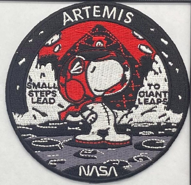 May include: Embroidered patch featuring Snoopy in a red and white spacesuit, with the word "ARTEMIS" at the top. The patch includes the NASA logo and the phrases "SMALL STEPS LEAD" and "TO GIANT LEAPS". The patch is round with a black border.