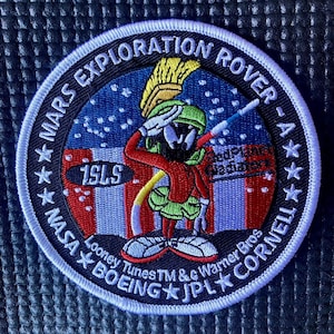 May include: A white embroidered patch with a blue background featuring a cartoon character saluting. The text "MARS EXPLORATION ROVER - A" is written in a circle around the character. The text "15LS" is written in a black oval below the character. The text "NASA BOEING JPL CORNELL" is written in a circle around the bottom of the patch. The text "Looney Tunes TM &c Warner Bros" is written below the text "NASA BOEING JPL CORNELL".
