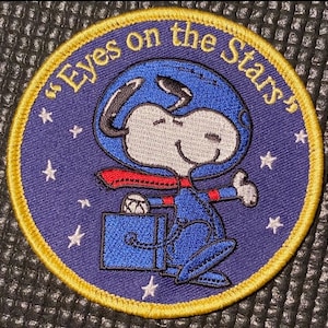 Op de afbeelding: Een geborduurde patch met Snoopy als astronaut met de tekst "Eyes on the Stars". De patch is blauw en geel.