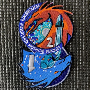 Puede incluir: Parche bordado con un diseño de dragón y cohete en tonos azules, naranjas y blancos. El parche incluye los nombres KIMBROUGH, MCARTHUR, HOSHIDE y PESQUET, junto con los números 1 y 2.