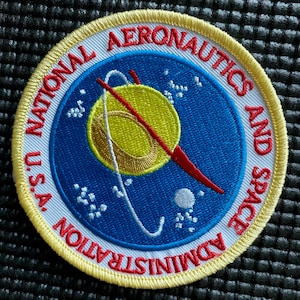 May include: A circular embroidered patch featuring the National Aeronautics and Space Administration U.S.A. logo. The design includes a blue background with a yellow planet, a red vector shape, a white orbital path, and white stars, all bordered by yellow.