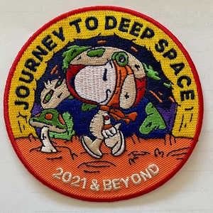 Puede incluir: Parche bordado circular con Snoopy con traje de astronauta, explorando un planeta con setas. El parche tiene el texto "JOURNEY TO DEEP SPACE" y "2021 & BEYOND" en amarillo y blanco.