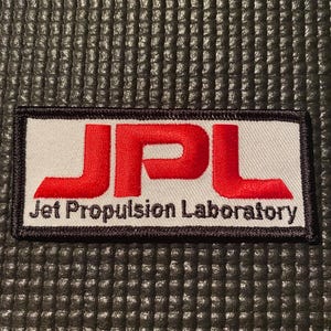 Op de afbeelding: Een rechthoekige patch met de rode letters "JPL" boven de zwarte woorden "Jet Propulsion Laboratory". De patch heeft een witte achtergrond en een zwarte rand. De achtergrond is een gestructureerd, donkergrijs oppervlak.