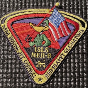 以下が含まれることがあります： 緑色の帽子をかぶって旗を持っている漫画の鴨が描かれた、黄色、赤、青の刺繍パッチ。パッチには「NASA Boeing JSF Commercial Red Planet Galactic」と書かれている