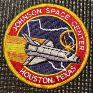 Può includere: Una toppa ricamata in rosso, bianco e giallo con il testo "Johnson Space Center Houston, Texas" e uno shuttle spaziale che vola sopra un contorno stilizzato dello stato del Texas.