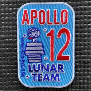 Peut inclure: Un écusson brodé bleu et blanc avec le texte "APOLLO 12 LUNAR TEAM" et un dessin animé d'un astronaute en combinaison spatiale. Le chiffre "12" est en rouge.