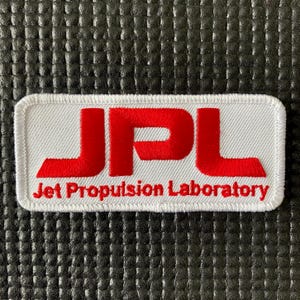 Può includere: Toppa rettangolare bianca con le lettere rosse "JPL" e la scritta "Jet Propulsion Laboratory" in rosso. La toppa ha un bordo bianco ed è su una superficie nera strutturata.
