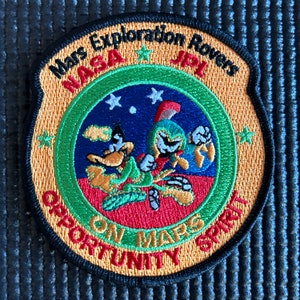 Puede incluir: Un parche bordado amarillo y verde con el texto "Mars Exploration Rovers NASA JPL Opportunity on Mars Spirit" y una imagen de dibujos animados de dos astronautas.