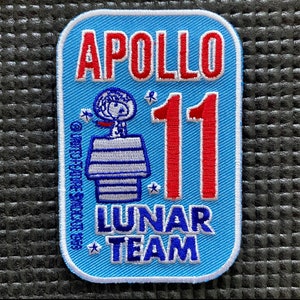 Pode incluir: Um remendo bordado azul com o texto "APOLLO 11 LUNAR TEAM" em letras brancas e vermelhas. Um astronauta de desenho animado está em pé em uma plataforma listrada branca e azul com estrelas ao redor.