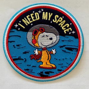 Può includere: Una toppa ricamata blu e bianca con un cane dei cartoni animati che indossa una tuta spaziale e un casco. Il testo "I NEED MY SPACE" è scritto intorno al cane.
