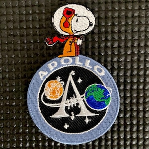 Könnte beinhalten: Gestickter Aufnäher mit Snoopy in einem Astronauten-Outfit, der auf einem blau-schwarzen kreisförmigen Aufnäher steht. Der Aufnäher trägt das Wort "APOLLO" und Bilder von Planeten und Sternen. Die Snoopy-Figur ist weiß, orange und rot.
