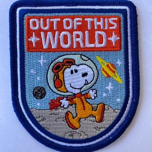 Puede incluir: Un parche bordado con Snoopy vestido de astronauta con un traje espacial azul y naranja. El parche tiene un borde azul y el texto "OUT OF THIS WORLD" en letras rojas y blancas.