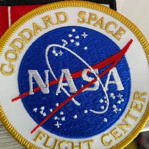 May include: A white embroidered patch with a blue NASA logo, red diagonal line, and gold lettering that reads "Goddard Space Flight Center".