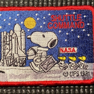 Könnte beinhalten: Ein gestickter Aufnäher in Rot, Weiß und Blau mit einer Zeichentrickfigur von Snoopy in einem Raumanzug, der vor einem Space Shuttle steht. Der Text "SHUTTLE COMMAND" und "NASA" ist ebenfalls auf dem Aufnäher gestickt.