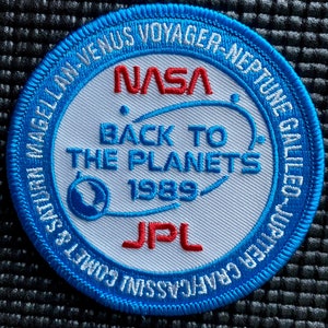 Puede incluir: Un parche de la NASA azul y blanco con el texto "Back to the Planets 1989" y los logotipos "NASA" y "JPL". El parche también incluye los nombres de las misiones espaciales: "Voyager-Neptune", "Galileo", "Jupiter Craft", "Cassini", "Comet's Encounter", "Magellan Venus" y "Ulysses".