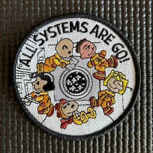 以下が含まれることがあります： 黒い縁取りのある白い刺繍パッチ。パッチには、洗濯機の周りを円形に囲む5人のキャラクターの漫画画像が描かれています。キャラクターの周りに「ALL SYSTEMS ARE GO!」というテキストが書かれています。