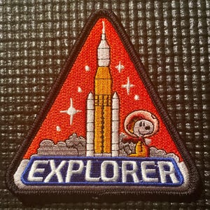 May include: Embroidered patch in a triangular shape with a red background, featuring a rocket, stars, and a cartoon astronaut. The word "EXPLORER" is displayed in a blue and white banner at the bottom.