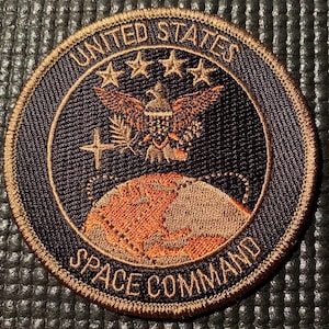 May include: A circular patch with the words "UNITED STATES SPACE COMMAND" in gold lettering. The patch features a gold border, a black background, and an eagle emblem above a globe.